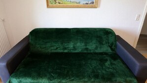 Een kamer met een donkerblauwe fauteuil, een groene velourse plaid en een lijst met een landschap erop.