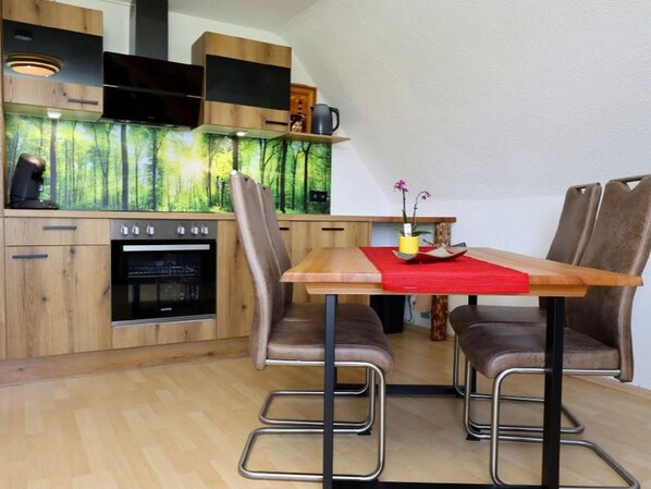 A modern kitchen with wooden cabinets, a built-in oven, and a dining area with a red tablecloth and metal chairs.