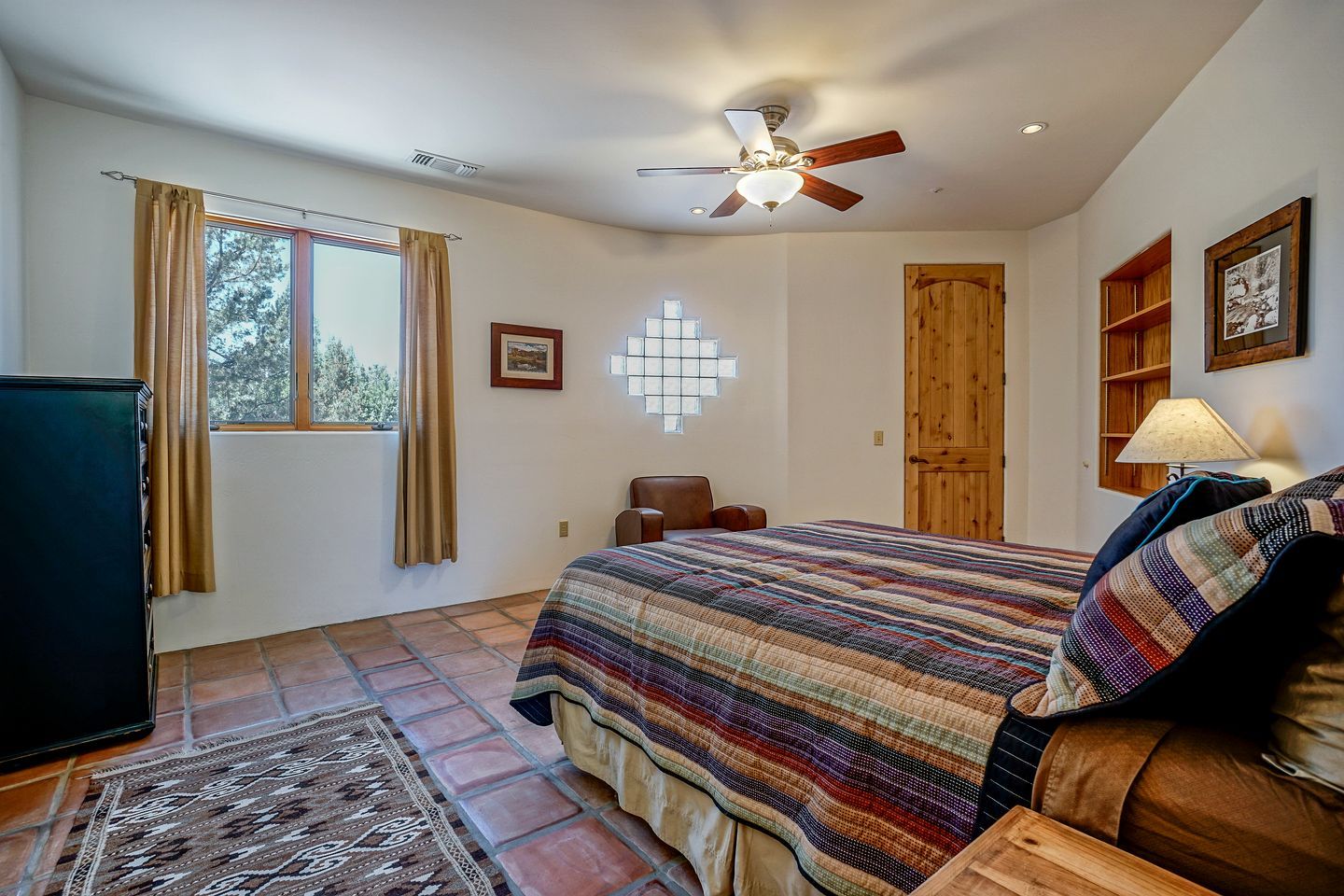 A bedroom with a bed, a ceiling fan, a window with curtains, a door, a wooden cabinet, and a television.