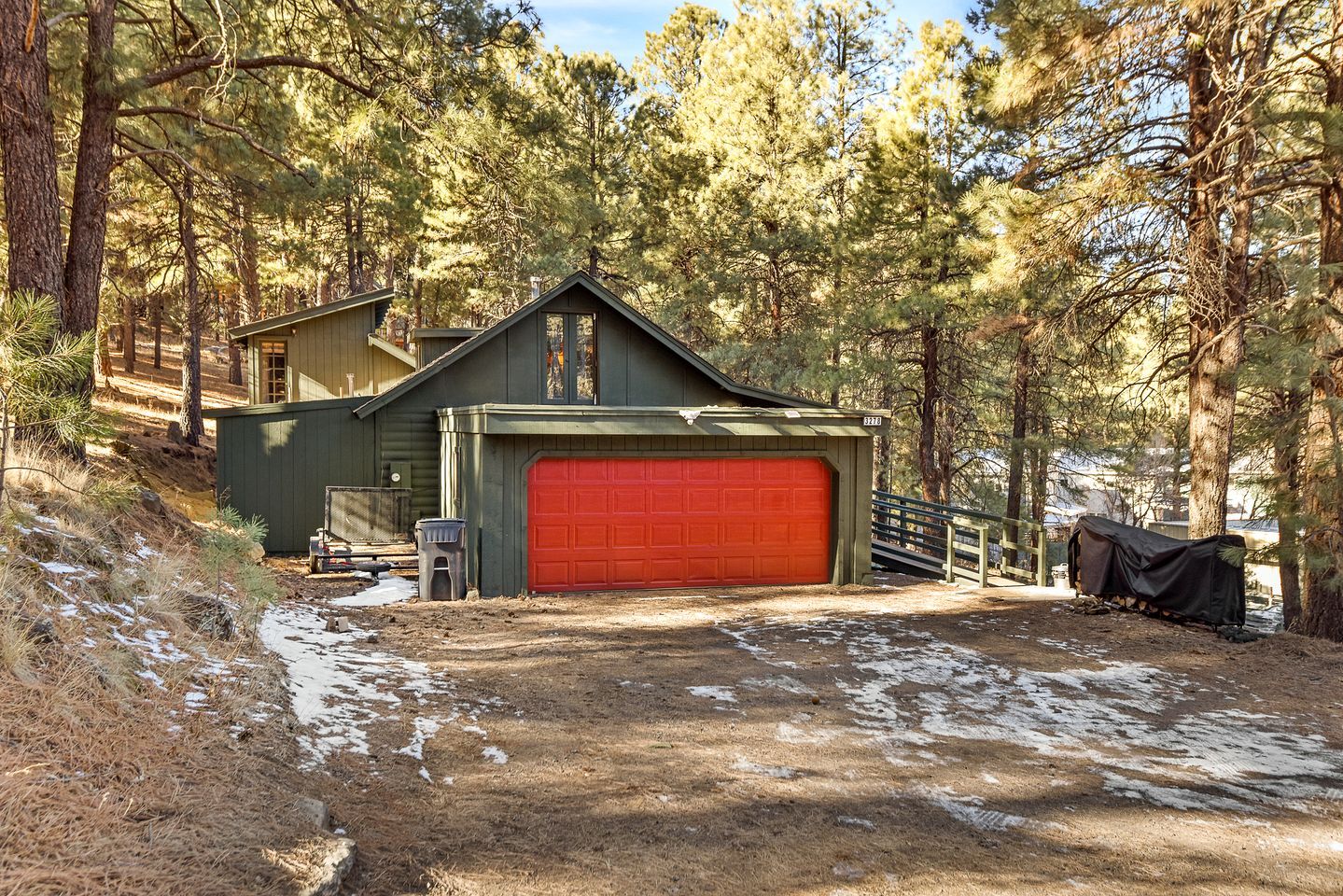 Une cabane à deux étages avec une porte de garage rouge, entourée d’arbres et donnant sur une allée en terre.