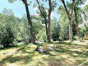 Una hamaca, dos sillas Adirondack, una mesa de picnic y un cobertizo pequeño en un área boscosa.