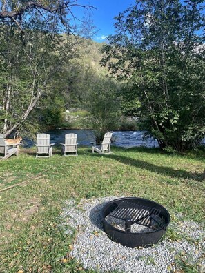 A lakeside outdoor seating area with Adirondack chairs, a fire pit, and a river in the background.