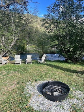 A lakeside outdoor seating area with Adirondack chairs, a fire pit, and a river in the background.
