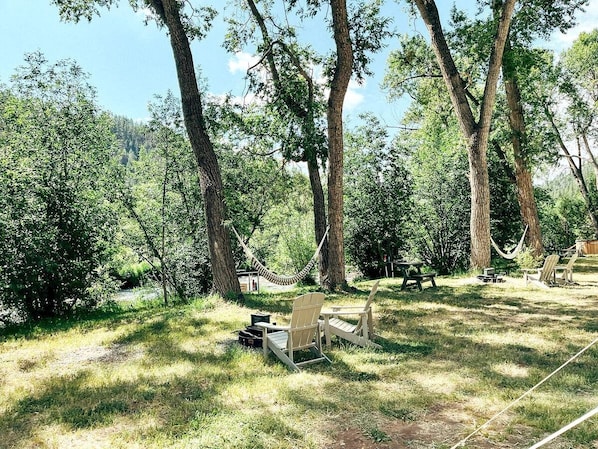 A hammock, two Adirondack chairs, a picnic table, and a small shed in a wooded area.