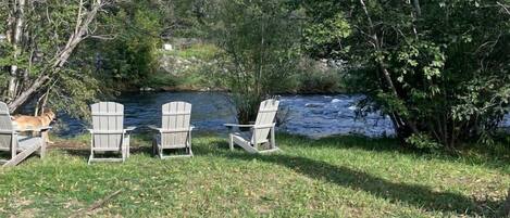 A lakeside outdoor seating area with Adirondack chairs, a fire pit, and a river in the background.