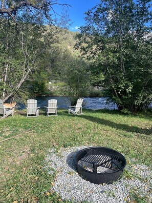 A lakeside outdoor seating area with Adirondack chairs, a fire pit, and a river in the background.