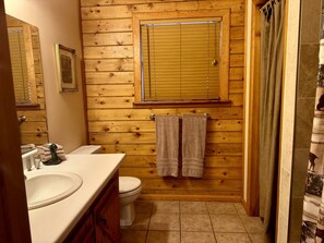 A bathroom with wooden paneling, a toilet, a sink, and a shower curtain. - Cozy Cabin with Indoor Jacuzzi Great for Couples in Arkansas (Eureka Springs)