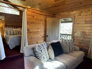 A cozy cabin interior with a sofa, a bed with a canopy, and a window with a view of greenery. - Cozy Cabin with Indoor Jacuzzi Great for Couples in Arkansas (Eureka Springs)