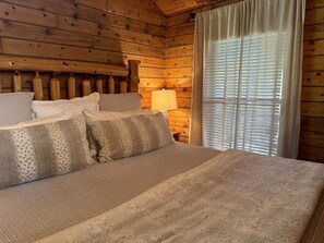 A wooden cabin-style room with a bed, pillows, a lamp, and a window with curtains. - Cozy Cabin with Indoor Jacuzzi Great for Couples in Arkansas (Eureka Springs)