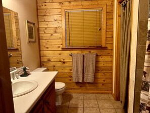 A bathroom with wooden paneling, a toilet, a sink, and a shower curtain. - Cozy Cabin with Indoor Jacuzzi Great for Couples in Arkansas (Eureka Springs)