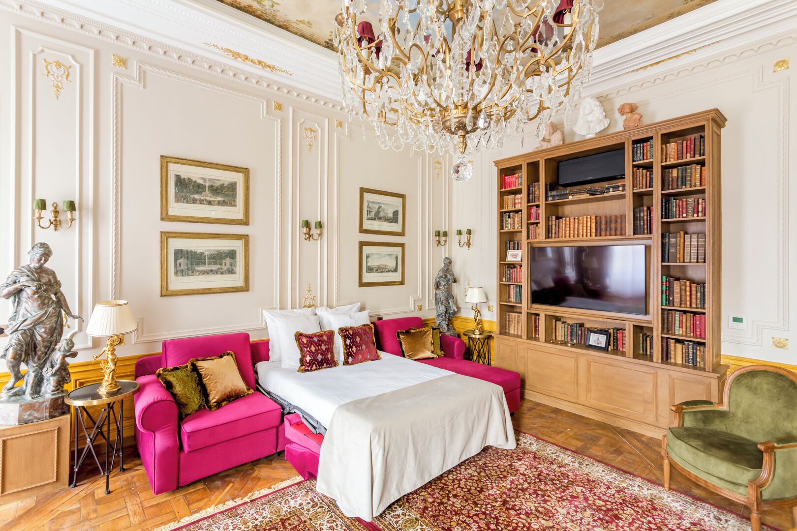 A luxurious bedroom with a large bed, a vibrant pink sofa, a wooden bookshelf, and a chandelier.