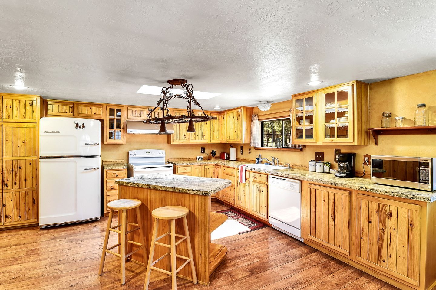 Cuina amb armaris de fusta, nevera, fogó, microones i un tamboret.