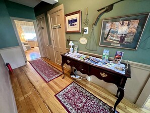 A hallway with wooden flooring, a washroom sign, a desk with papers, a framed picture, and a wall-mounted item. - Unique Cottage with Great Surroundings in Sunderland, Vermont (Sunderland)
