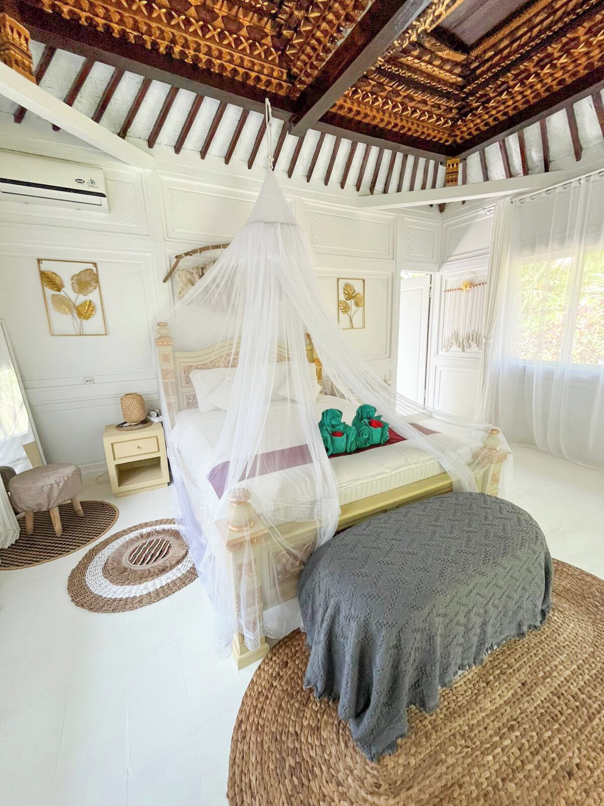 A bedroom with a canopy bed, a bedside table, a nightstand, a small stool, and a woven rug.