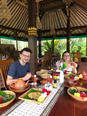 A traditional wooden dining area with a thatched roof, featuring a table set with local dishes, a teapot, and a woven basket.