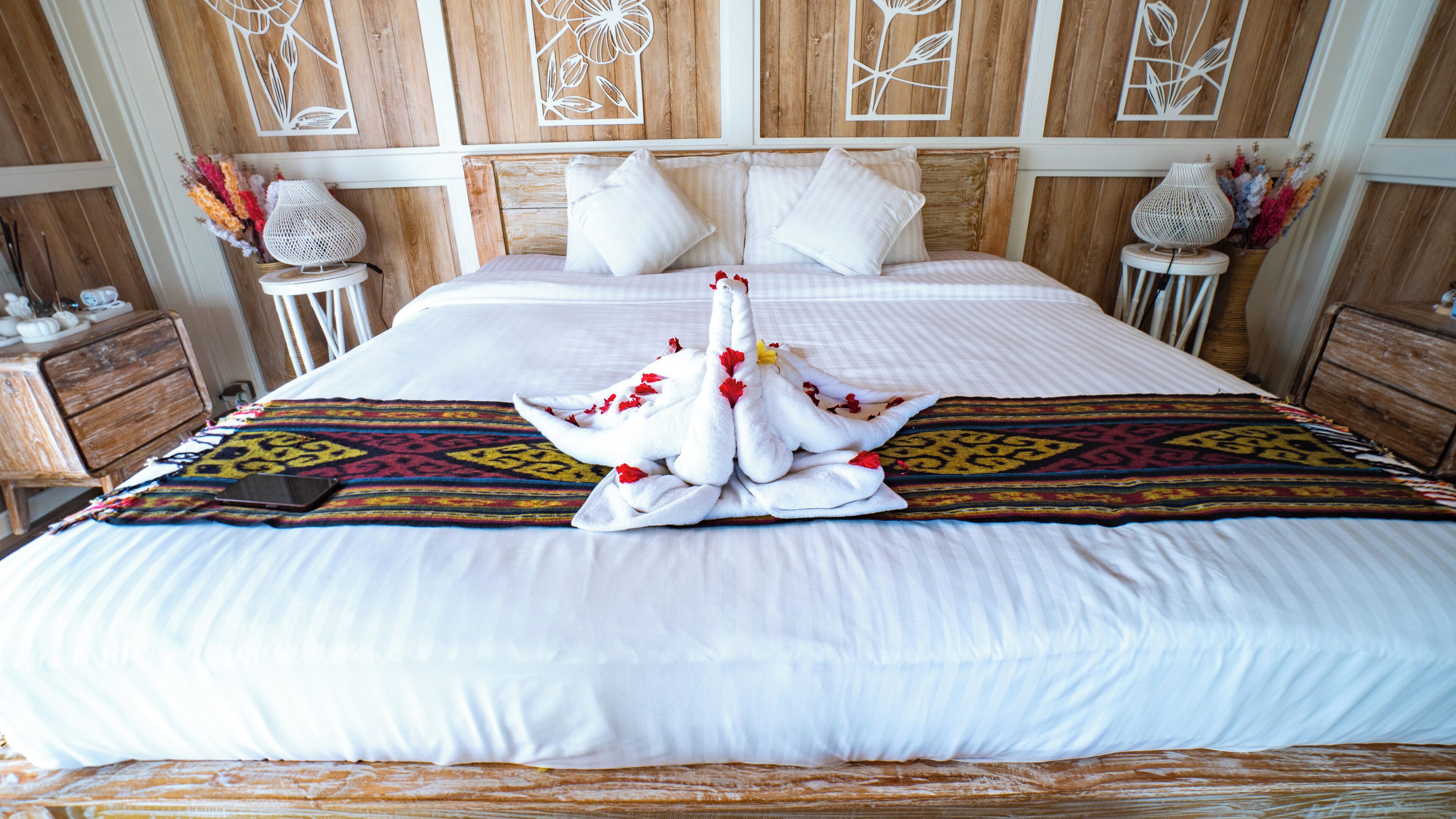 A neatly made bed with a decorative towel arrangement in the center, a patterned throw blanket, and wooden headboard panels with floral designs.