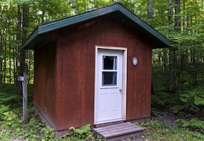 A small wooden cabin with a white door and a window, surrounded by dense forest.