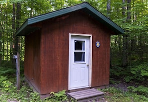 Una pequeña cabaña de madera con puerta blanca y ventana, rodeada de un bosque denso.