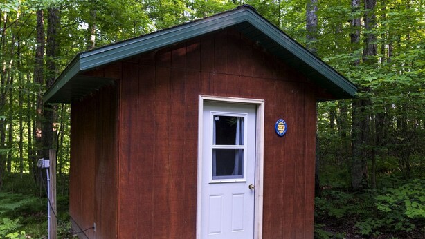 A small wooden cabin with a white door and a window, surrounded by dense forest.