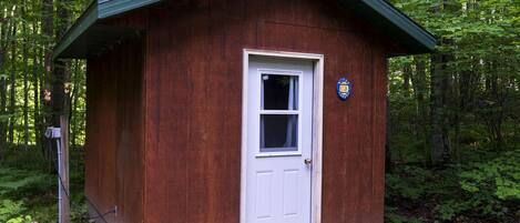 Una pequeña cabaña de madera con puerta blanca y ventana, rodeada de un bosque denso.