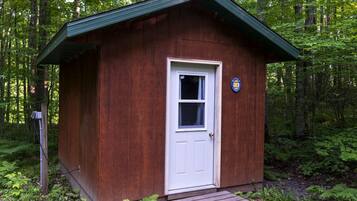 Une petite cabane en bois, avec une porte blanche et une fenêtre, entourée d’une forêt dense.