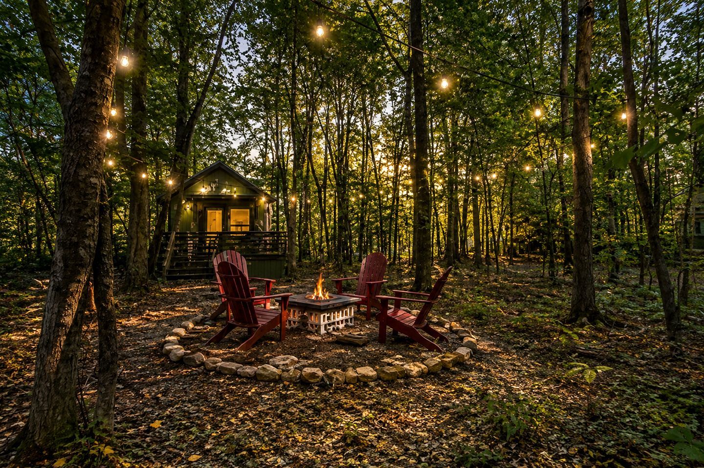 A cabin surrounded by trees with a fire pit and chairs in the center.