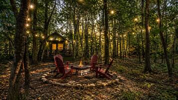 A cabin surrounded by trees with a fire pit and chairs in the center.
