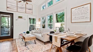 A spacious living room with a sofa, coffee table, and dining table. There are large windows, a ceiling fan, and artwork on the walls. - Luxurious Tiny Cabin with Hot-Tub and Fire Pit in Tennessee (Monteagle)