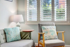 A room with two chairs, one with a patterned cushion and another with a floral cushion, a small round table with a lamp, and a window with blinds.