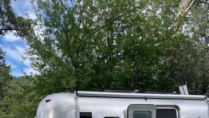 Ein silberner Airstream-Wohnwagen mit Schiebetür, zwei Adirondack-Stühlen, einem kleinen gelben Tisch und einer Wiese mit Bäumen im Hintergrund.
