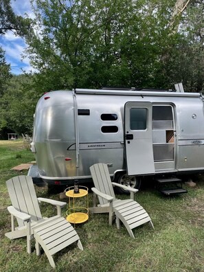 A silver Airstream trailer with a sliding door, two Adirondack chairs, a small yellow table, and a grassy area with trees in the background. - Unforgettable Glamping Escapes in this Cool Airstream on the Pecos River, New Mexico (Pecos)