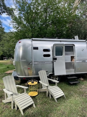 Una caravana Airstream plateada con puerta corredera, dos sillas Adirondack, una mesa amarilla pequeña y un césped con árboles al fondo.