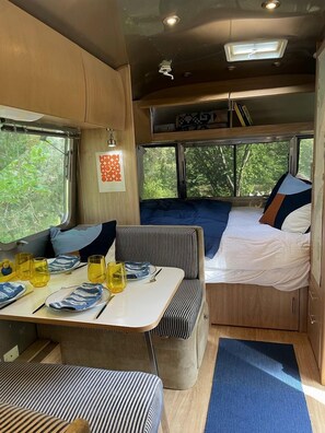 Interior compacto de una caravana con comedor, cama y ventana con vista a follaje.