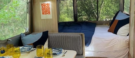 A compact RV interior with a dining area, a bed, and a window with a view of greenery.
