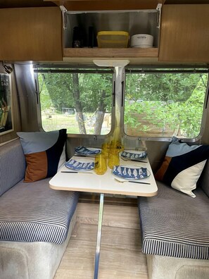 A compact RV interior with a dining table, two cushioned seating areas, and a window offering a view of greenery. - Unforgettable Glamping Escapes in this Cool Airstream on the Pecos River, New Mexico (Pecos)