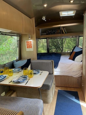 A compact RV interior with a dining area, a bed, and a window with a view of greenery.