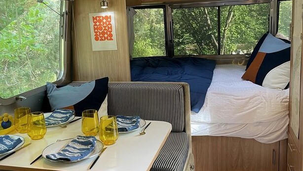 Ein kompakter Wohnraum in einem Wohnmobil mit Essbereich, einem Bett und einem Fenster mit Blick auf Grünflächen.
