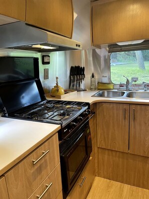 Cocina pequeña y funcional con estufa, horno, lavamanos, armarios de madera y ventana con vistas al paisaje.