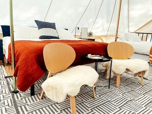 A room with a bed, two chairs, a small table, and a white furry stool.