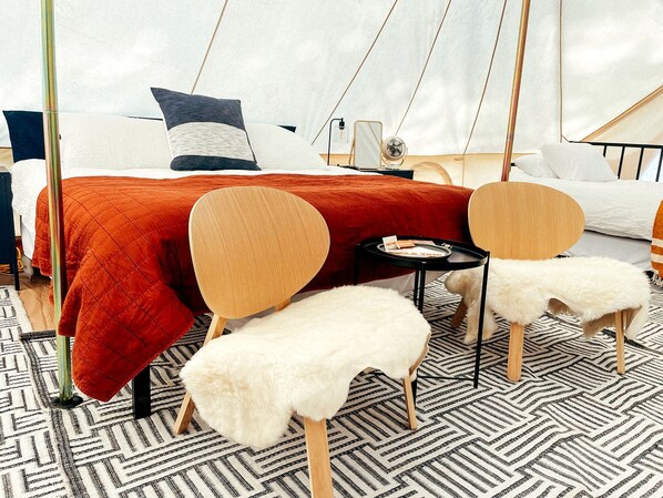 A room with a bed, two chairs, a small table, and a white furry stool.