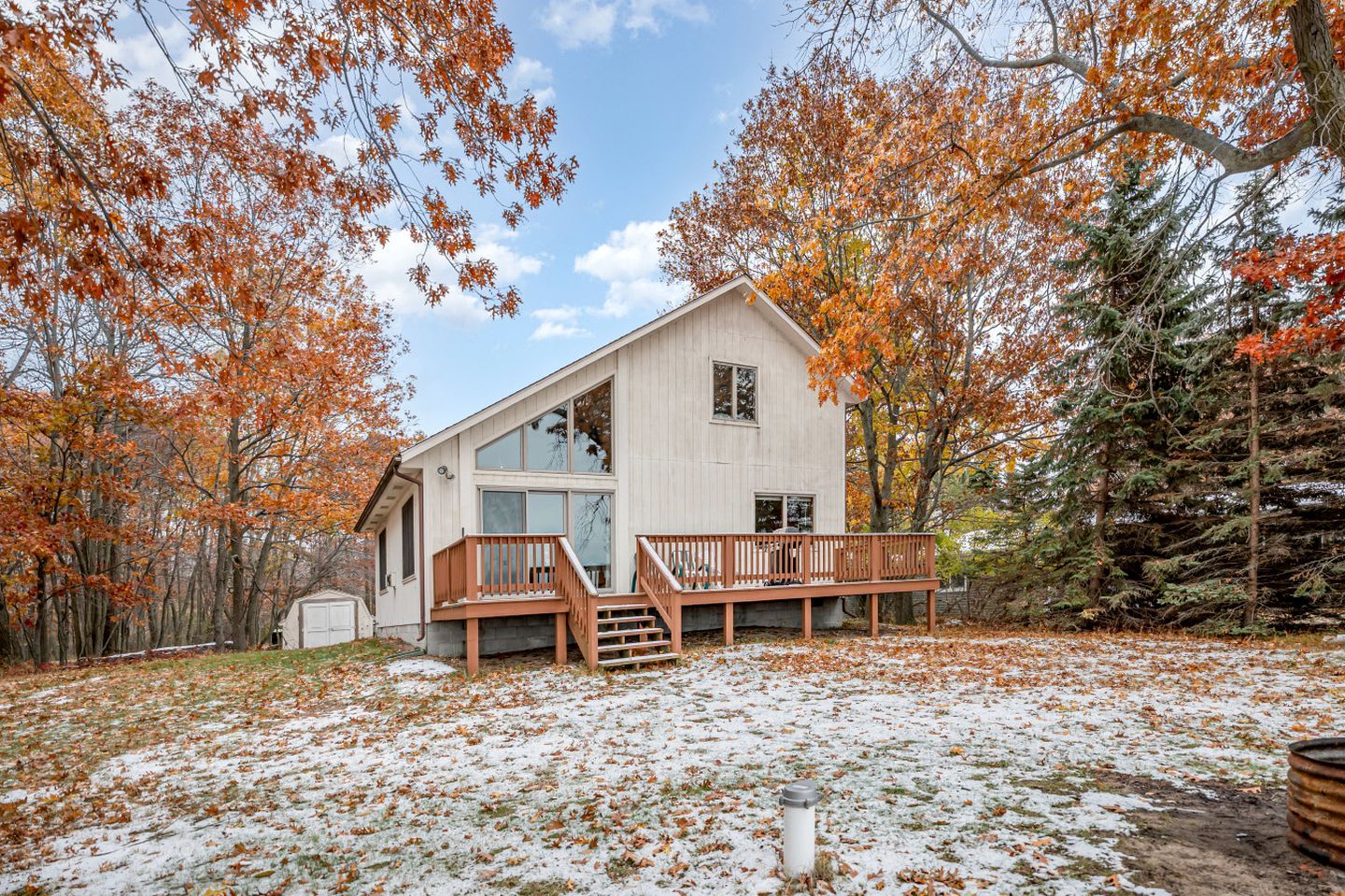 A two-story cabin with a wooden deck and large windows, surrounded by trees with autumn foliage.