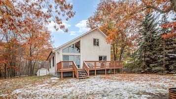 A two-story cabin with a wooden deck and large windows, surrounded by trees with autumn foliage.
