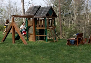 A wooden play structure with a swing set and climbing elements, surrounded by grass and trees. - Incredible Riverfront Cabin Surrounded by Trees for an Unforgettable Escape in Nature in Blue Ridge, Georgia (Blue Ridge)