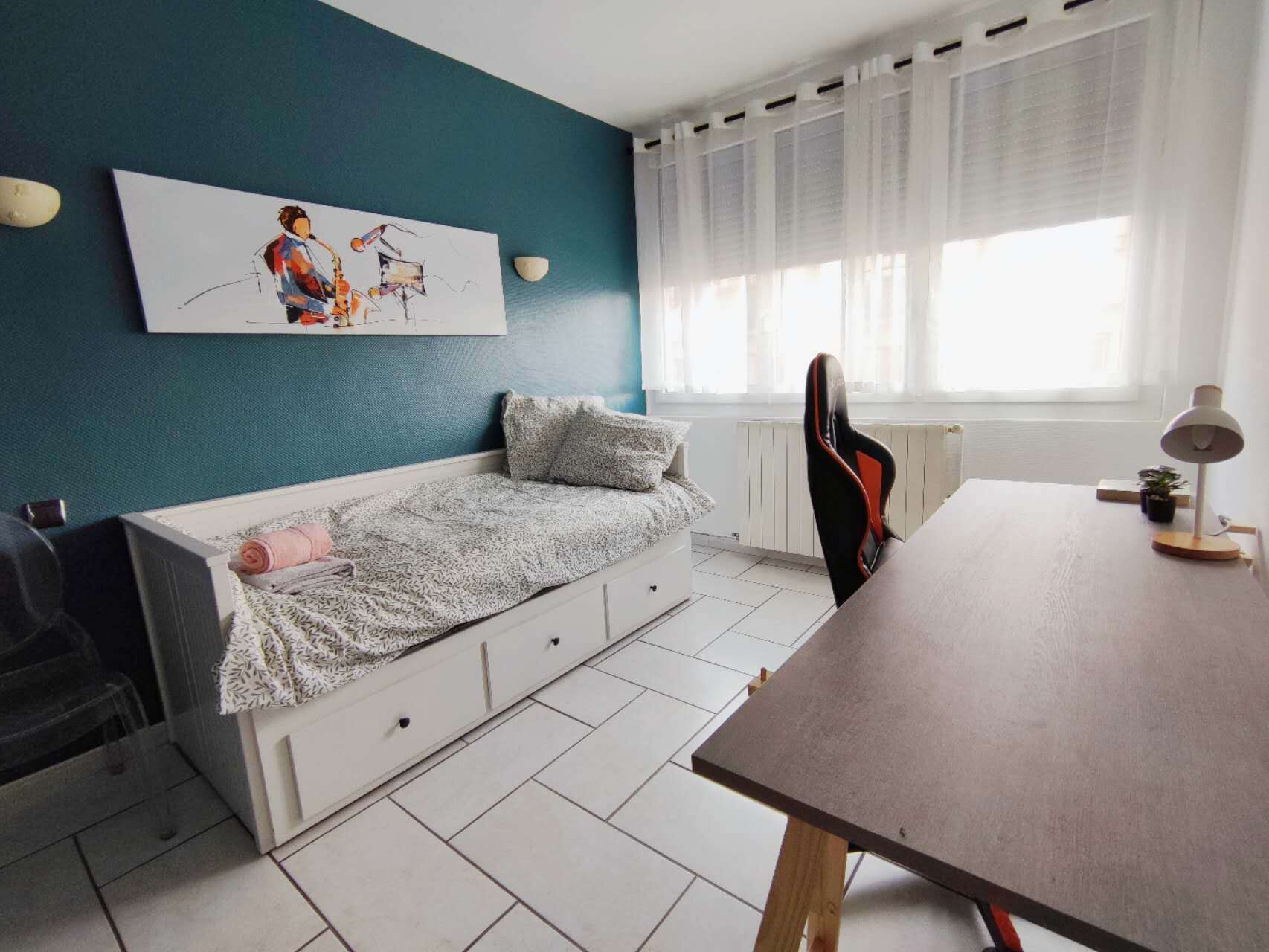 Une chambre avec un lit, un bureau et une chaise. Il y a une fenêtre avec des rideaux, un radiateur et un tableau accroché au mur.