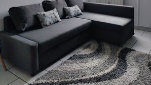 Ein modernes Wohnzimmer mit einem grauen Ecksofa, zwei Zierkissen, einer schwarzen Wanduhr, einem großen Fenster mit Jalousien und einem schwarz-weißen Teppich.