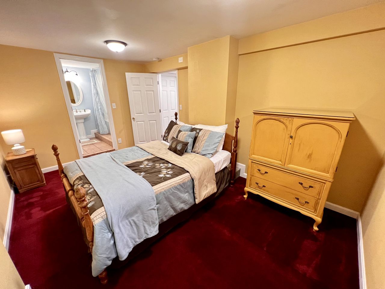 A bedroom with a bed, a wooden dresser, a nightstand, and a bathroom visible through an open door.