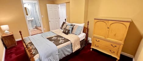 Un dormitorio con una cama, un aparador de madera, una mesita de noche y un baño visible a través de una puerta abierta.