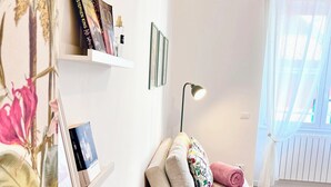 Ein gemütliches Wohnzimmer mit einer Couch, einem kleinen Hocker, einer Lampe, einem Bücherregal mit Büchern und einem Fenster mit durchsichtigen Vorhängen.