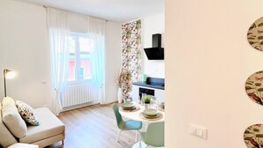 Ein modernes Wohnzimmer mit einer Couch, einem Esstisch und Stühlen. Ein Fenster mit Vorhängen, eine Pflanze und eine Küchenzeile mit einer schwarzen Dunstabzugshaube.