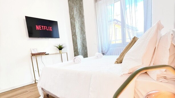 Ein modernes Hotelzimmer mit Bett, einem Fernseher, der 'NETFLIX' anzeigt, einem Nachttisch mit Pflanze und einer Lampe.